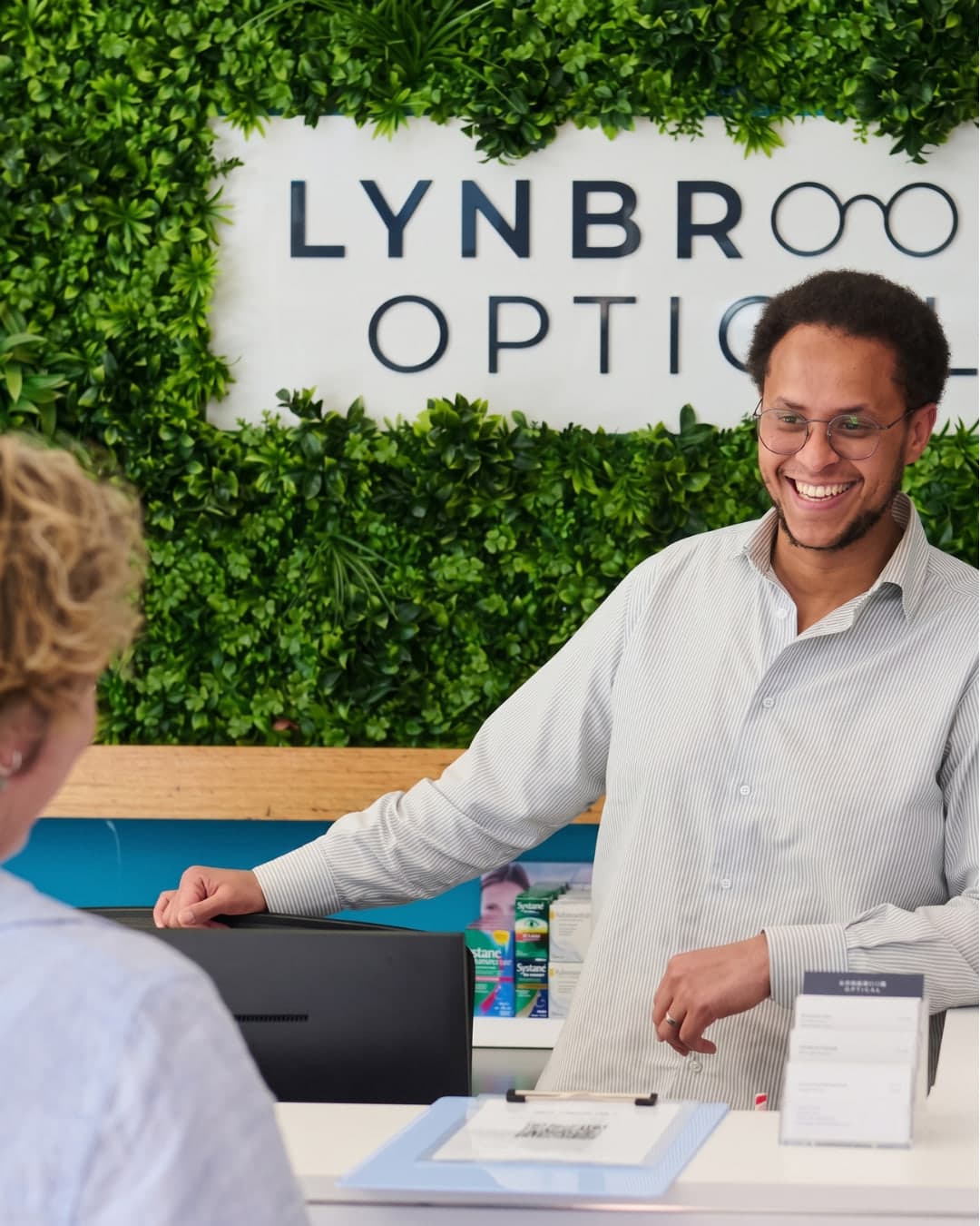 An optometrist at the front desk of Lynbrook Optical, smiling mid-conversation with a patient in front of the practice's signage